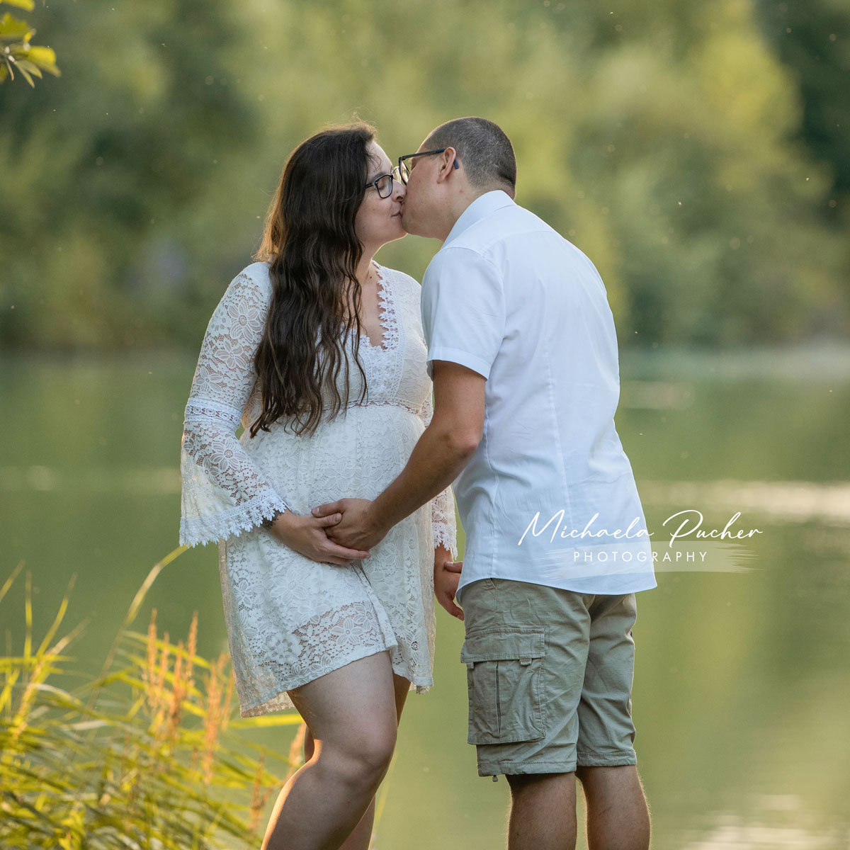 Schwangere Frau in weißem Spitzenkleid steht am See und küsst ihren Partner, der ihre Hände auf dem Babybauch hält.